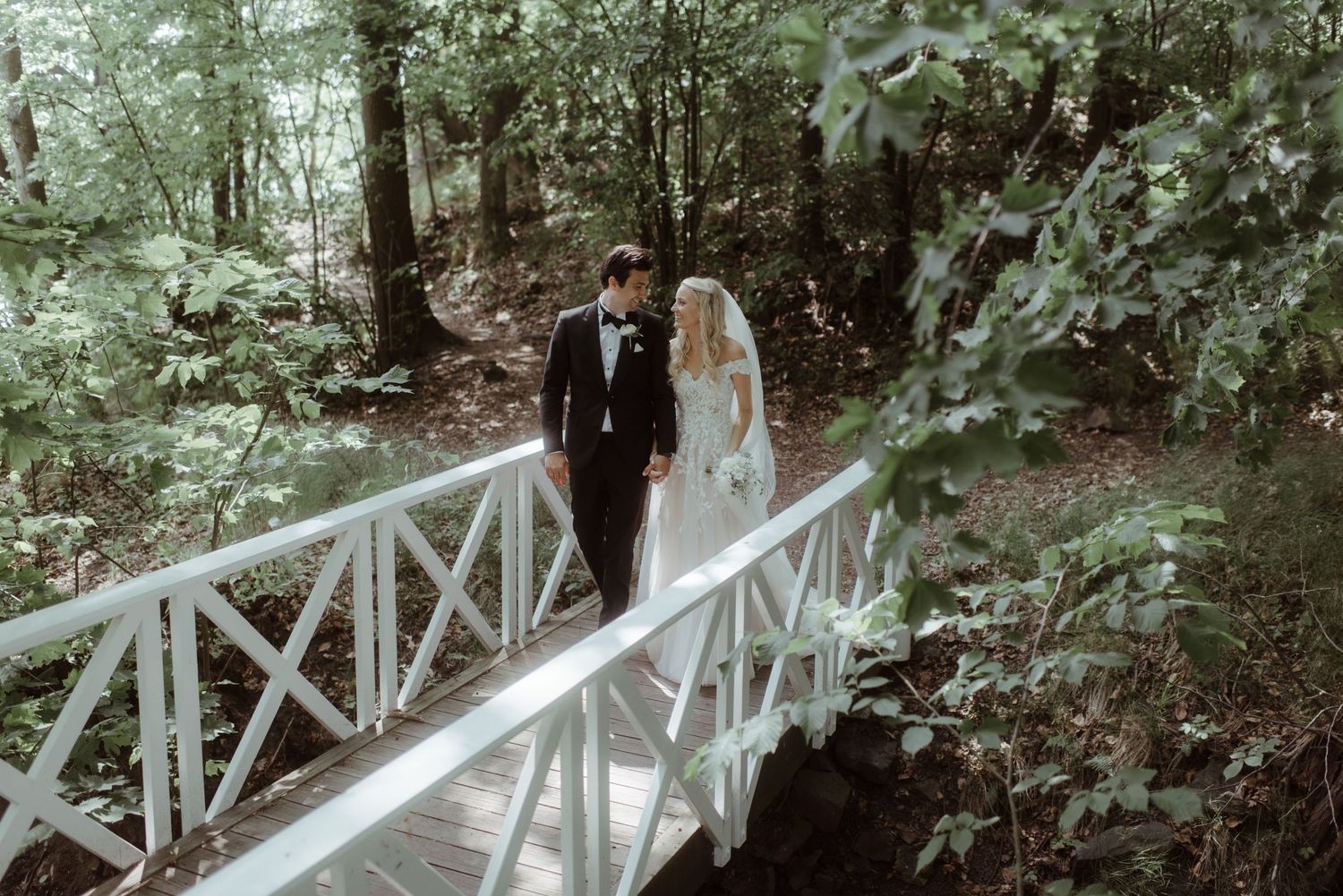 A couple in wedding attire stand on a white wooden bridge surrounded by lush forest greenery.