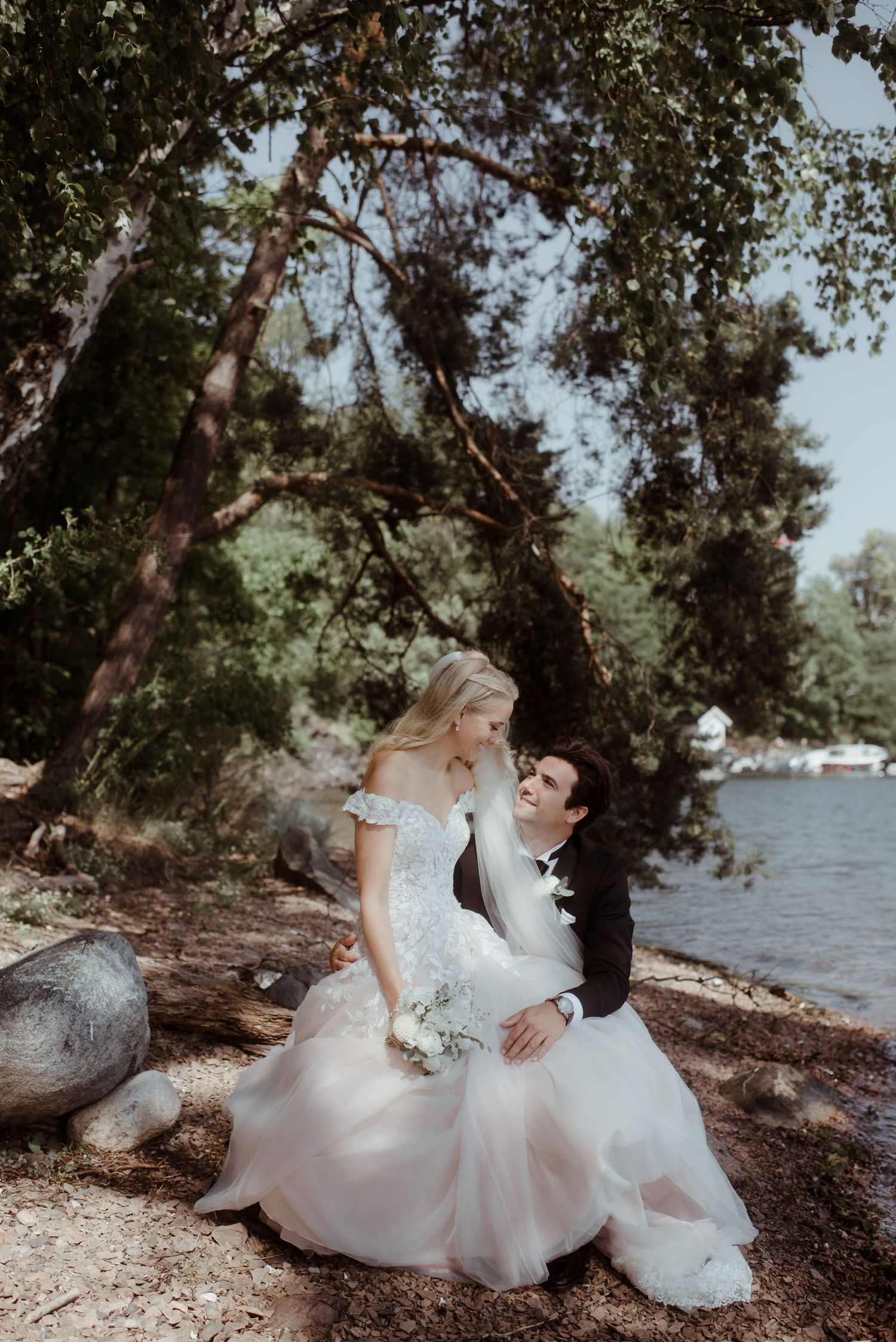 Newlyweds in formal attire sit together on a rocky shoreline under overhanging trees.