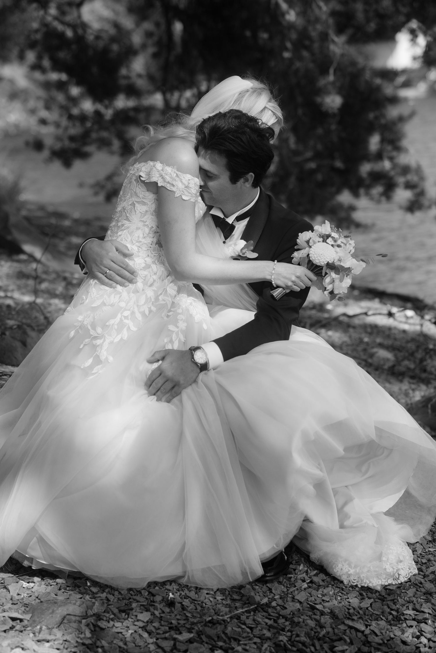 A romantic black and white wedding photo of a couple embracing in a lace dress and tuxedo outdoors.