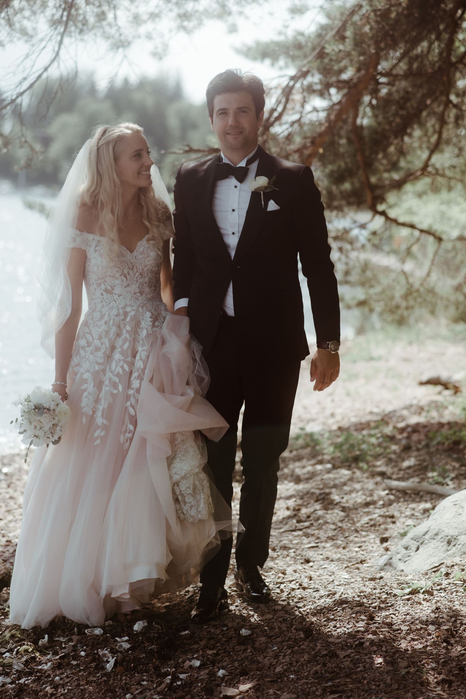 A wedding couple walks together along a forested path in formal attire.