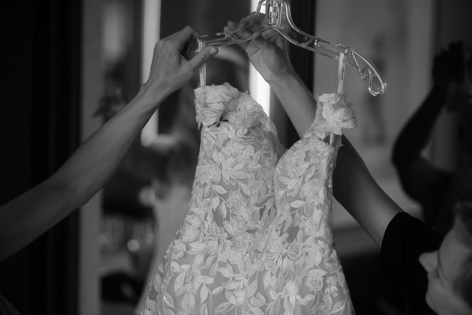 Multiple hands hold up an ornate white lace wedding dress in black and white.