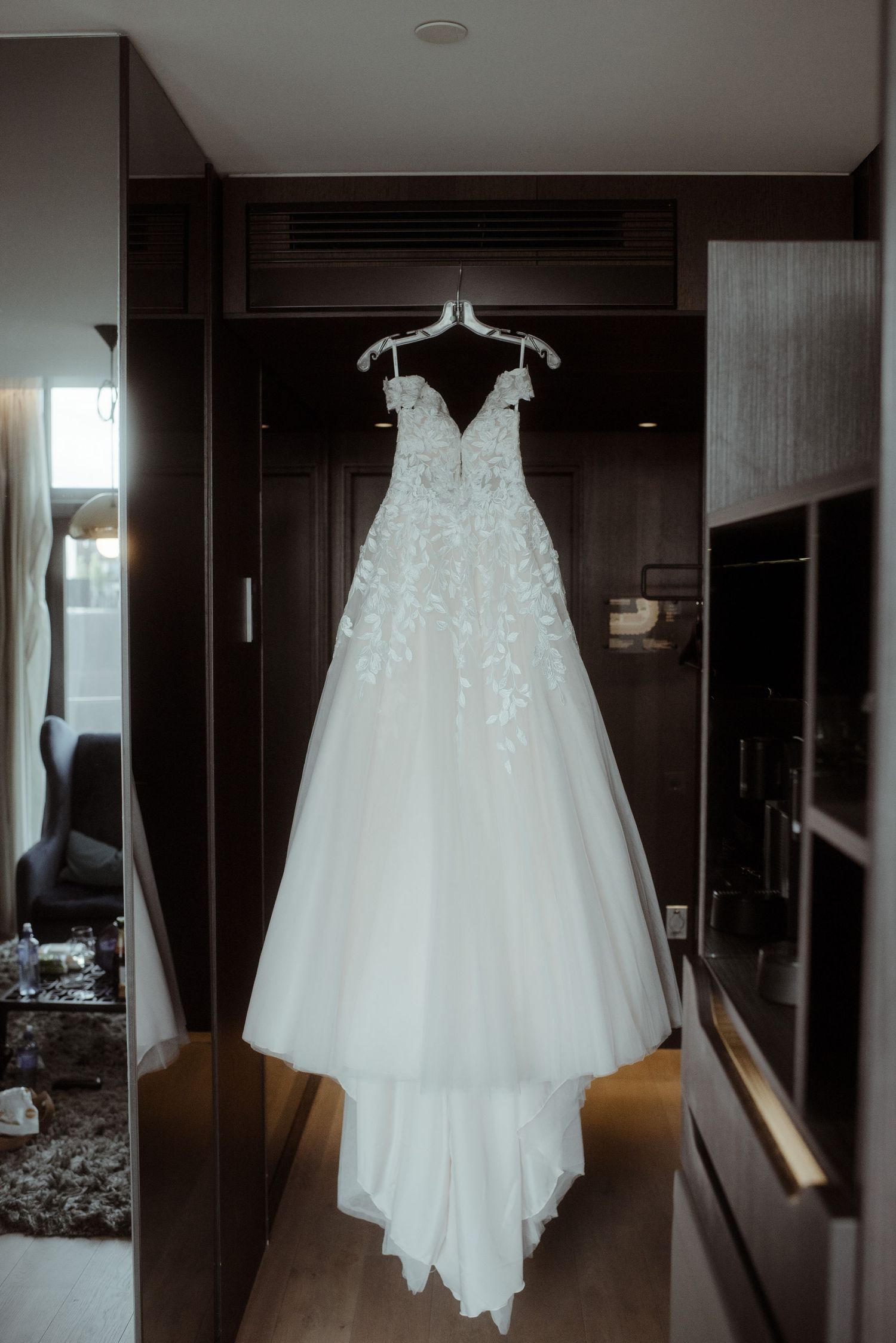 Elegant white lace wedding gown hanging in front of dark wooden wardrobe.