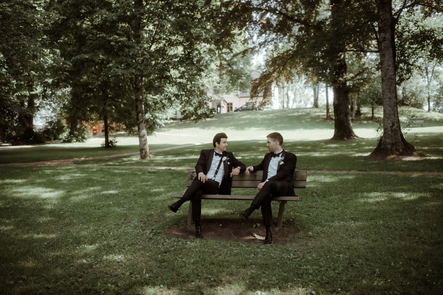Two people in tuxedos sharing a casual conversation on a park bench.
