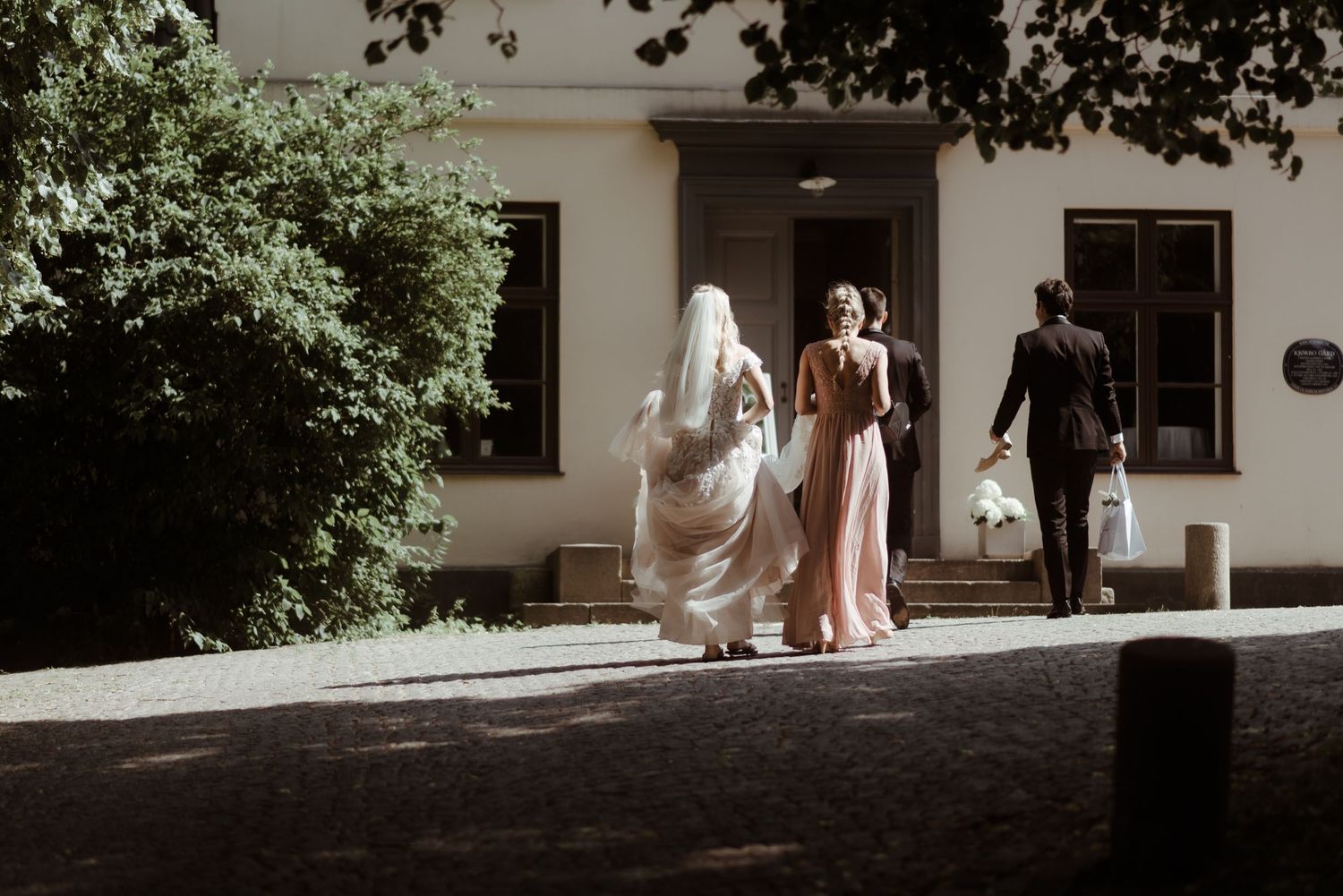 Wedding party members walk along a sunlit exterior pathway of a white building.