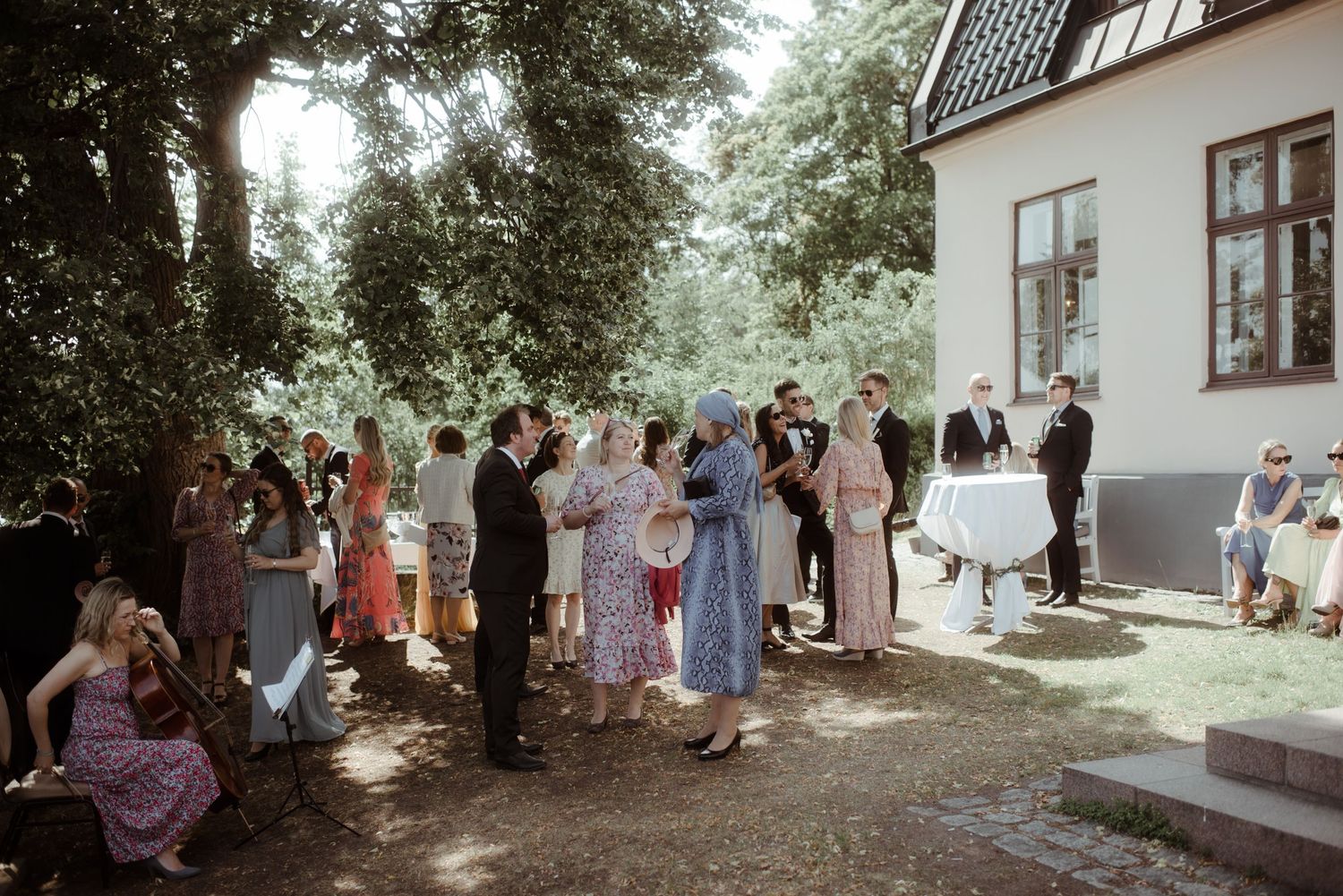 Wedding guests mingle in shaded outdoor garden area beside white house.