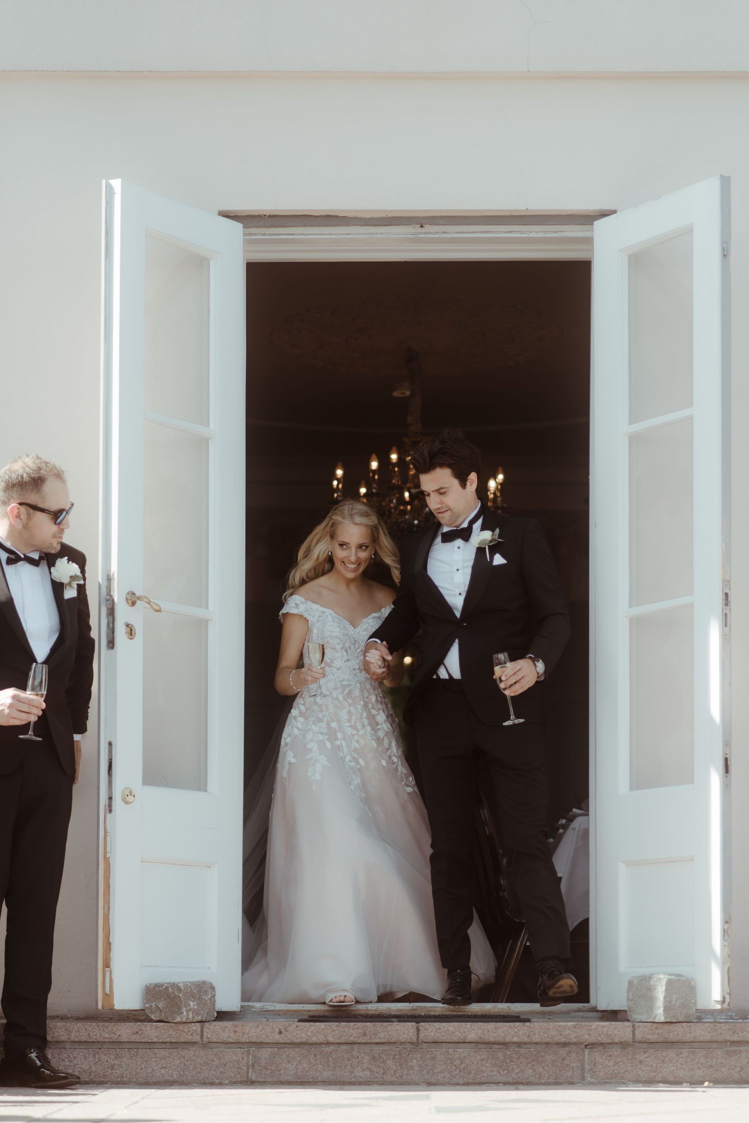 A couple exits through white double doors in formal wedding attire while guests hold champagne glasses.