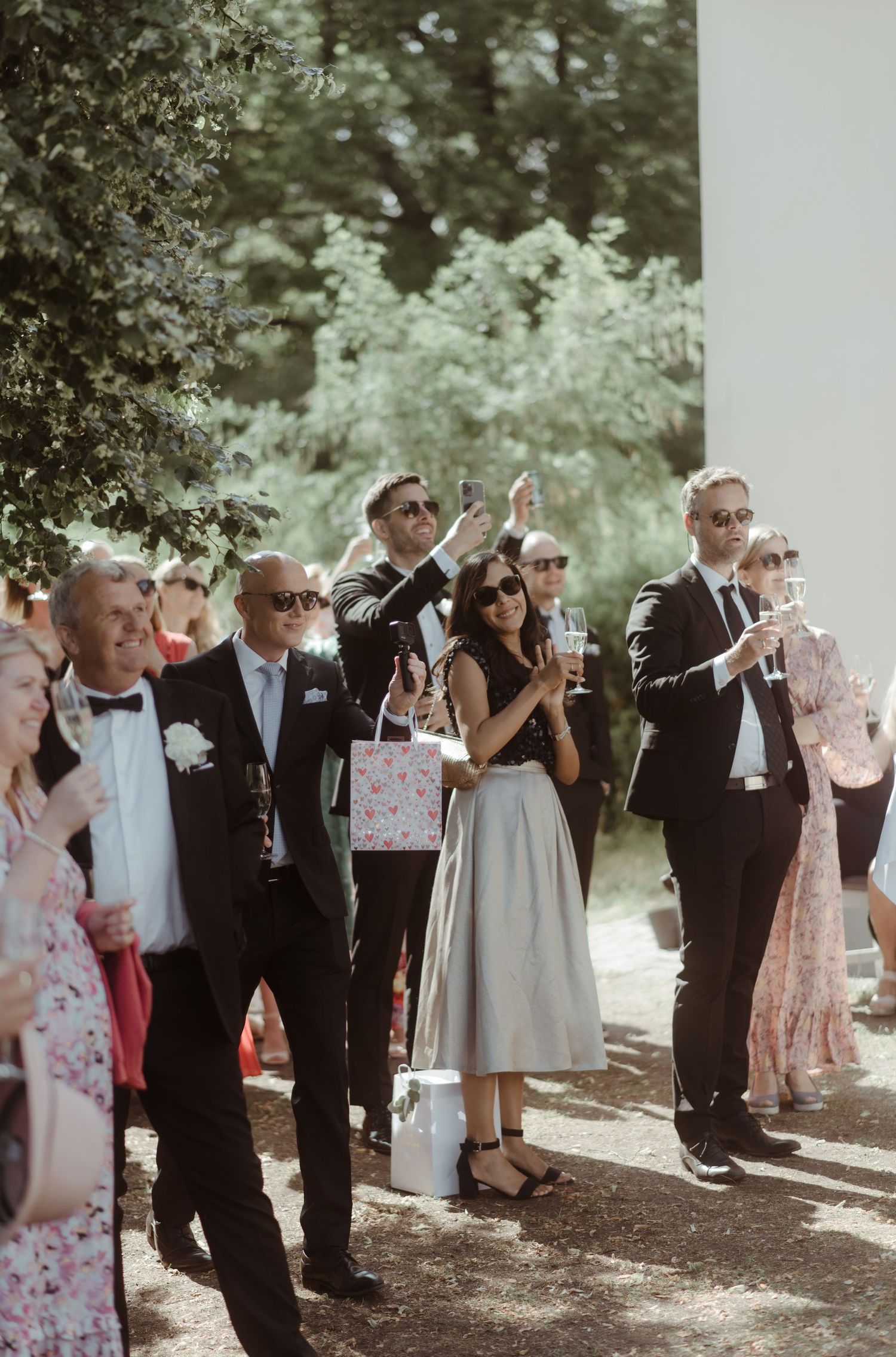 Wedding guests in formal attire toast and celebrate outdoors during a summer reception.