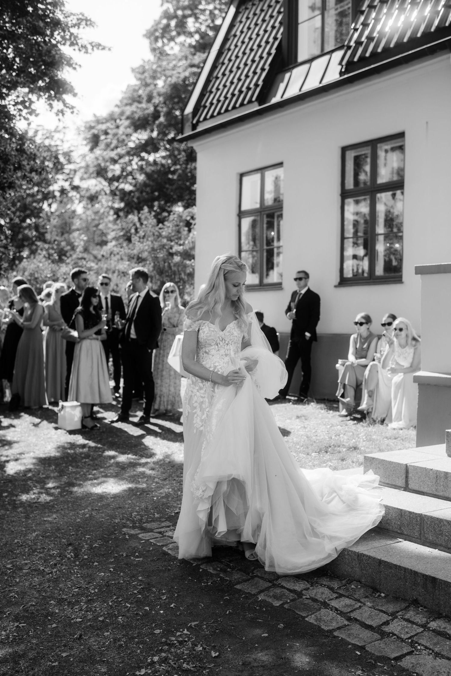 Guests gather outside a white house to watch the formal wedding proceedings in black and white.