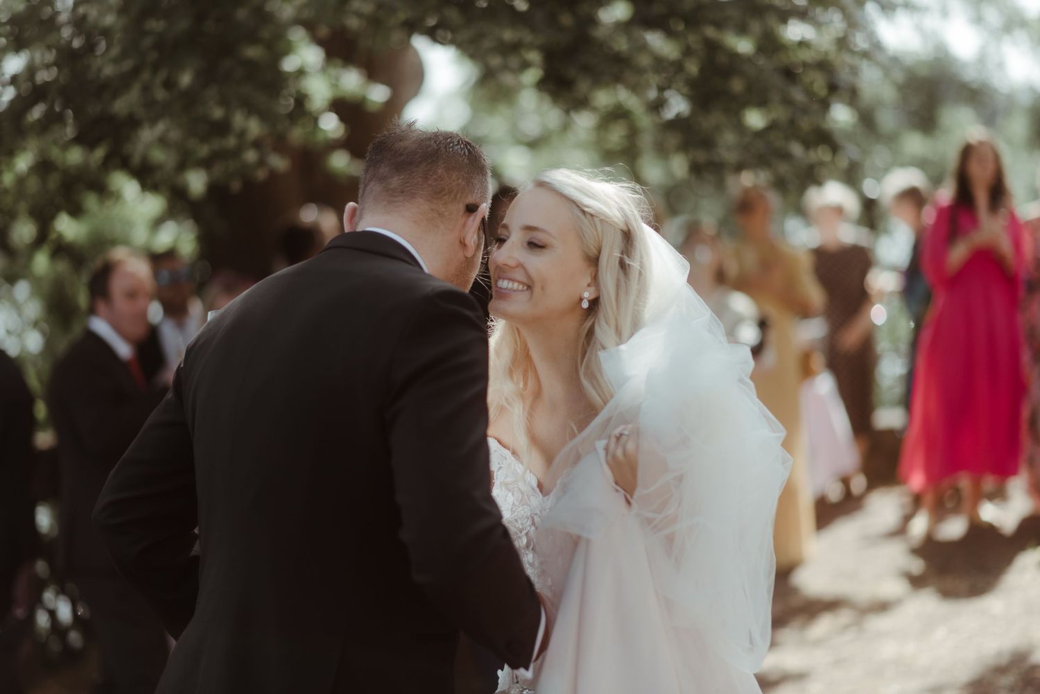 A romantic wedding moment captured in soft sunlight as guests look on.