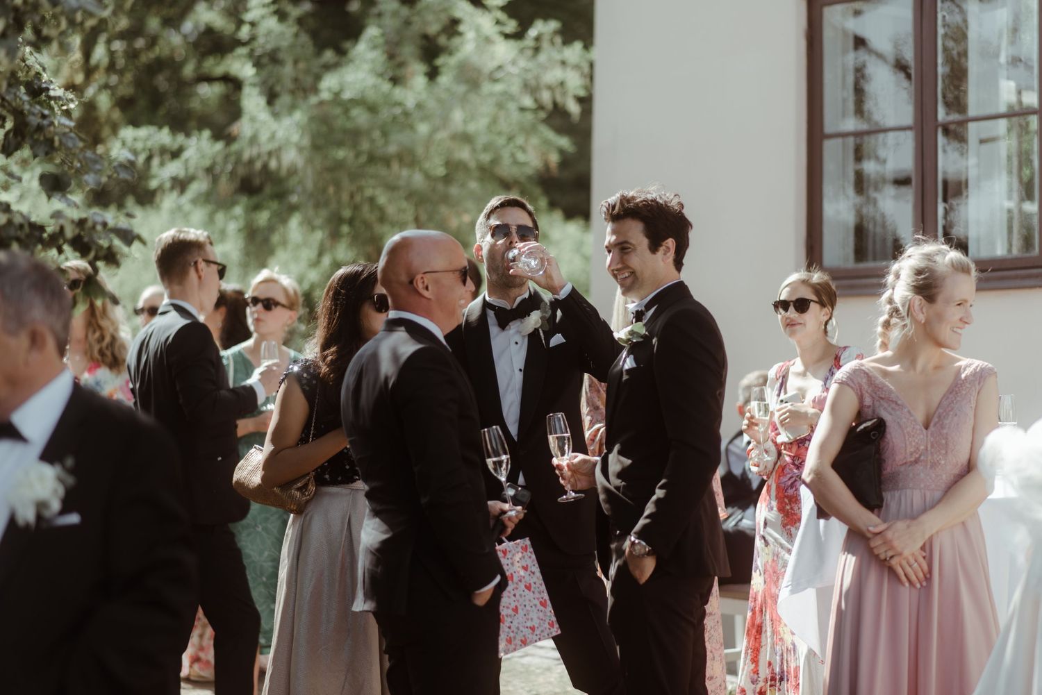 Guests in formal attire gather at an outdoor wedding celebration, mingling and chatting in natural daylight.