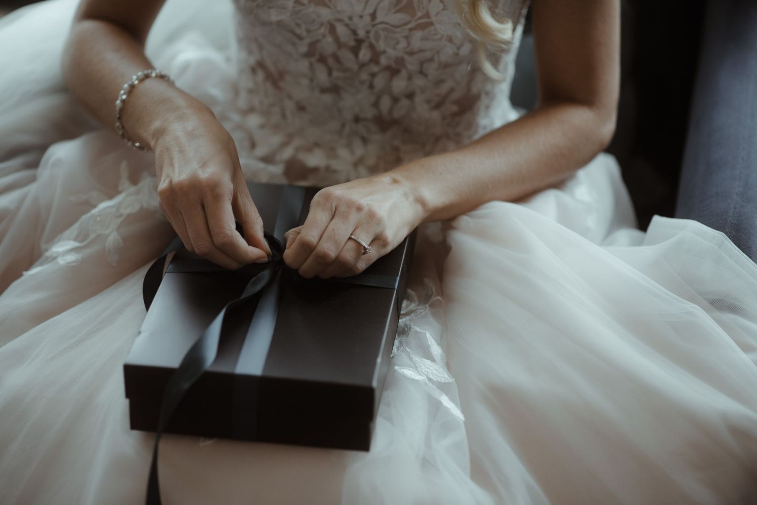 Bride in white dress opening a small dark box while seated.