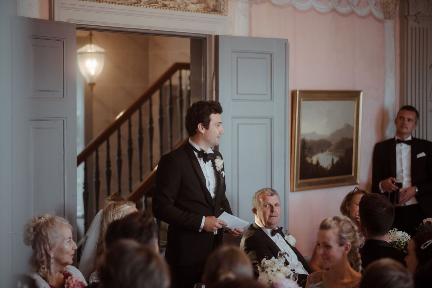 Wedding reception speech in an elegant room with traditional decor and staircase.