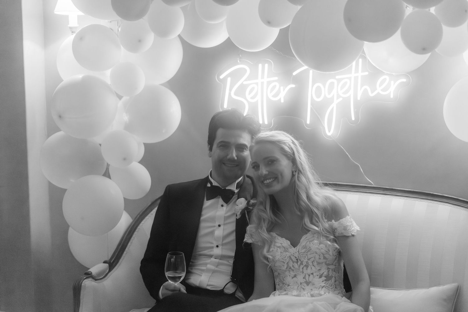 A newlywed couple sits beneath a 'Better Together' neon sign and white balloon arch at their wedding reception.