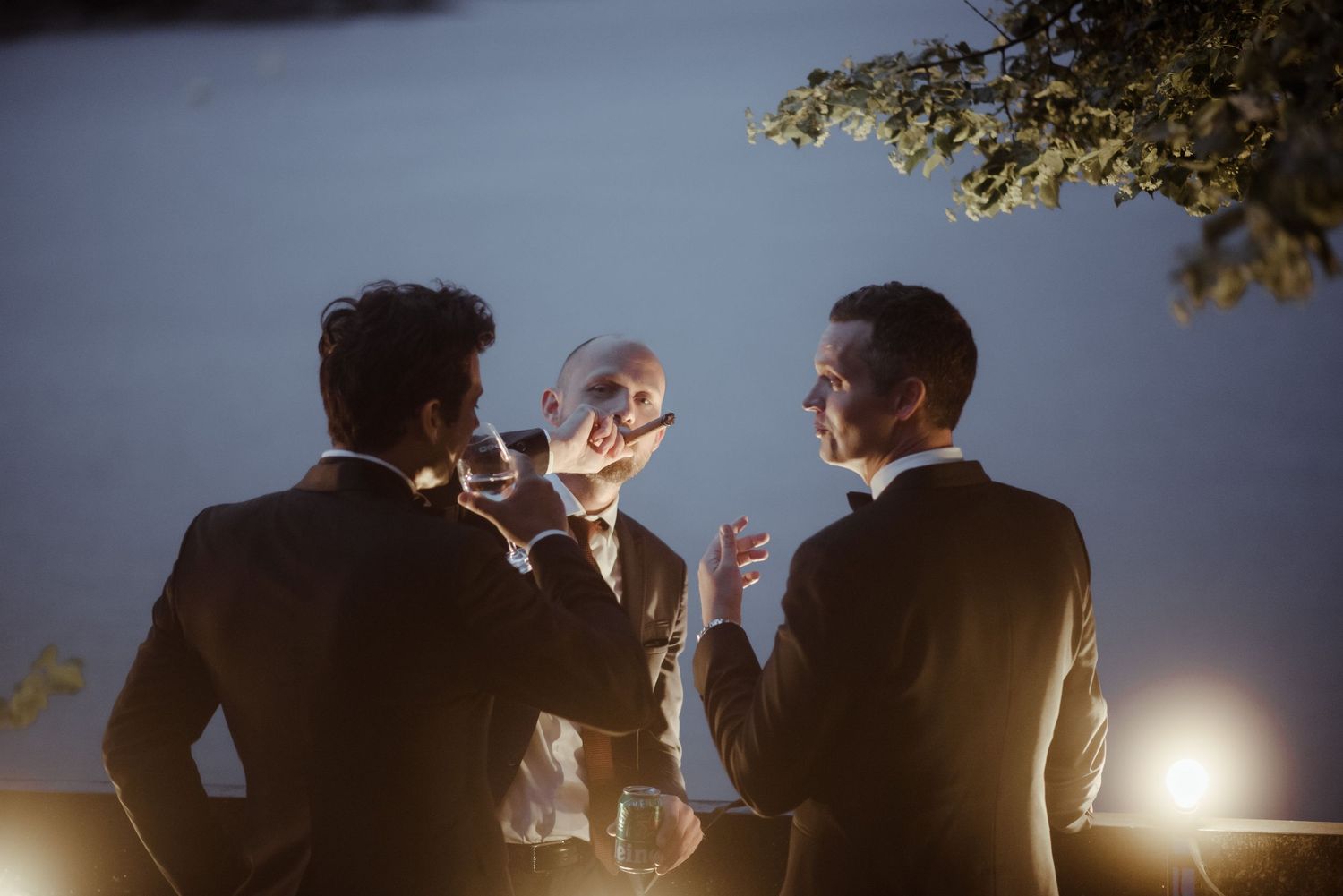 Silhouettes of guests enjoying drinks at dusk during an outdoor wedding celebration.