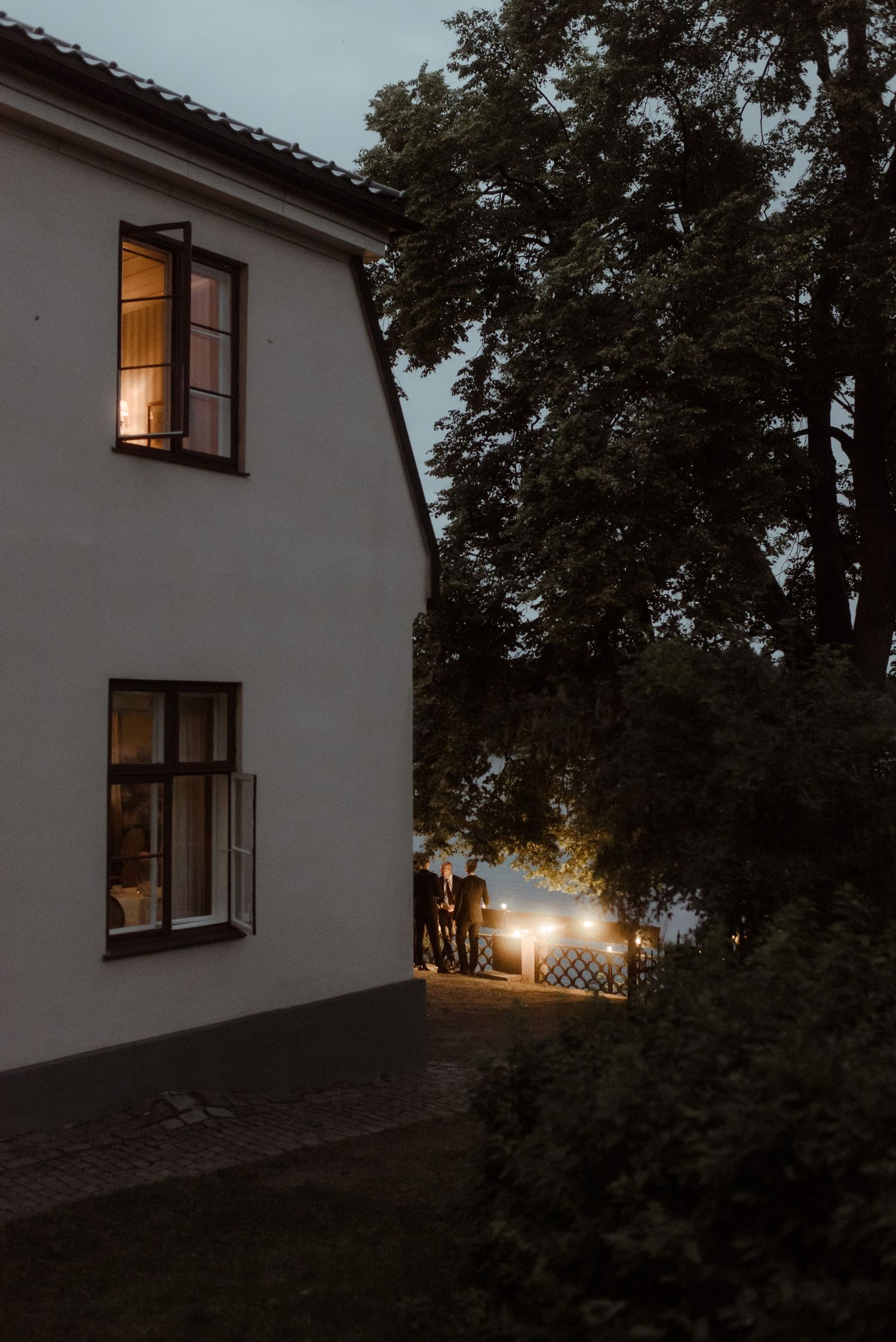 A warmly lit house exterior at dusk with guests visible through illuminated windows.