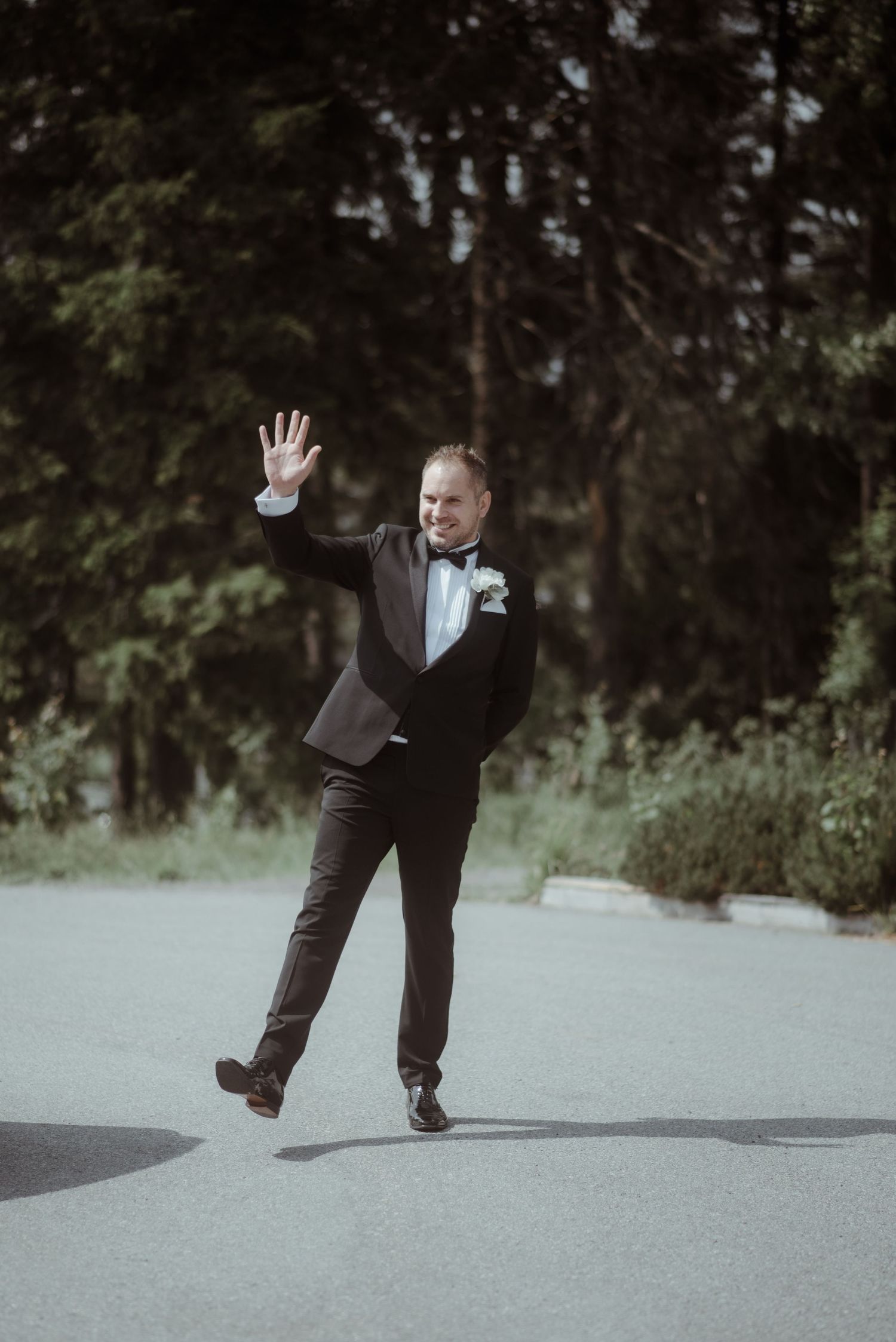 A groom in a classic tuxedo strikes a playful pose outdoors.