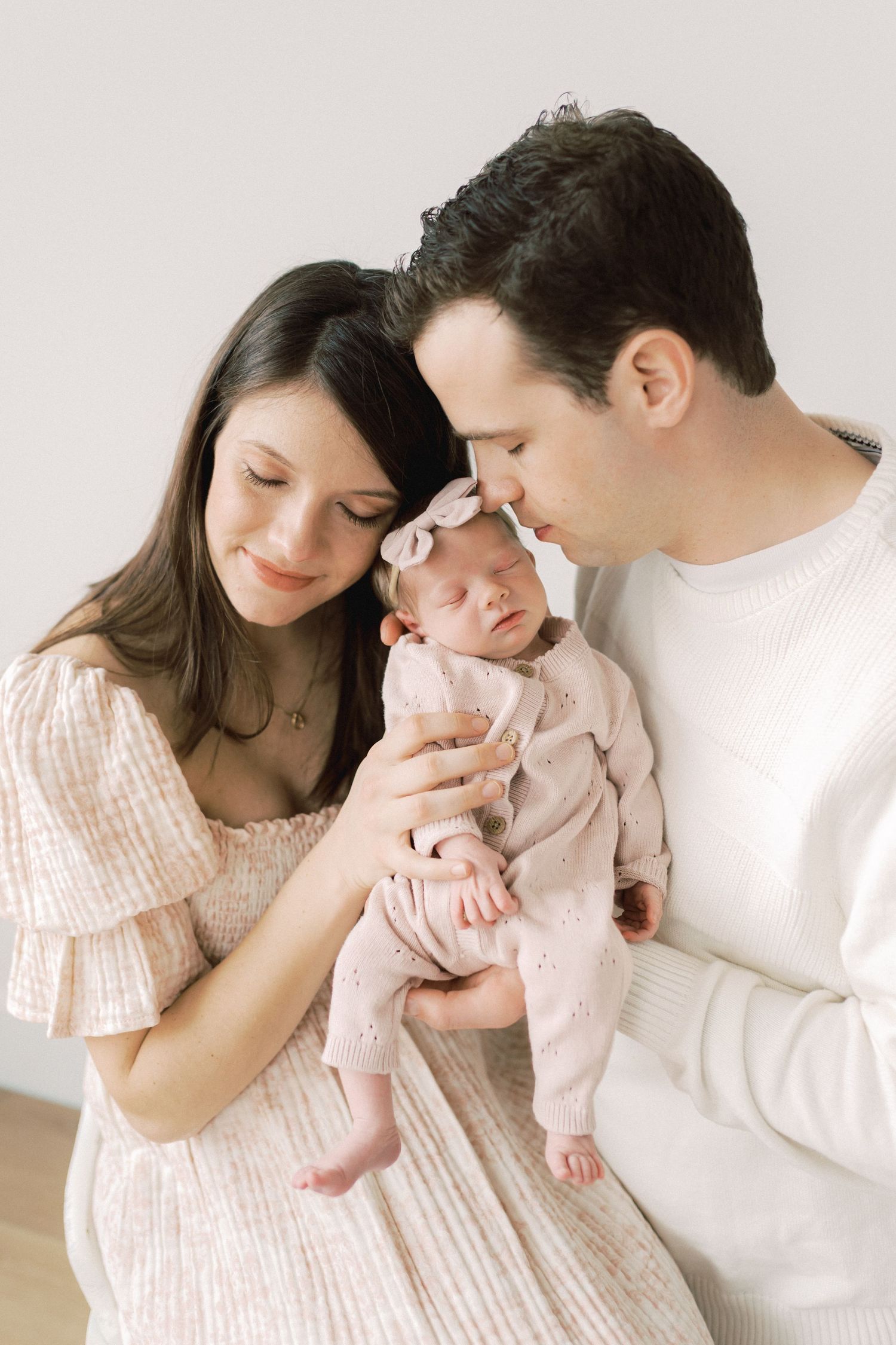 A tender family moment in soft pink and white tones as parents gaze lovingly at their sleeping newborn baby.