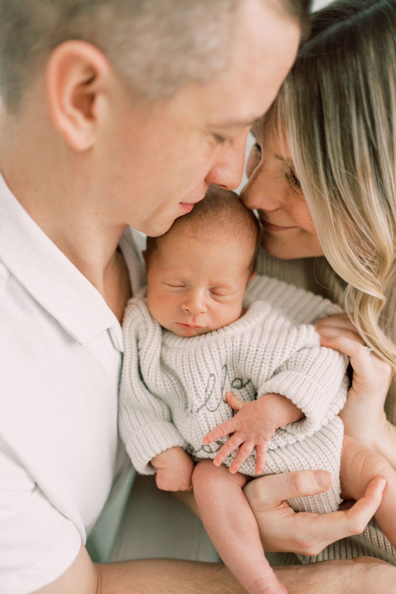 Intimate family portrait as parents lovingly embrace their sleeping newborn baby wearing a soft white sweater.