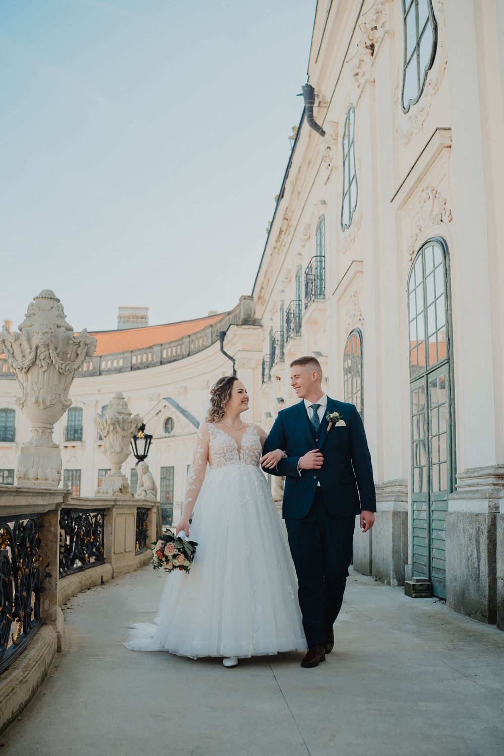 A wedding couple walks together along an elegant balcony with ornate architecture and tall windows in the background.