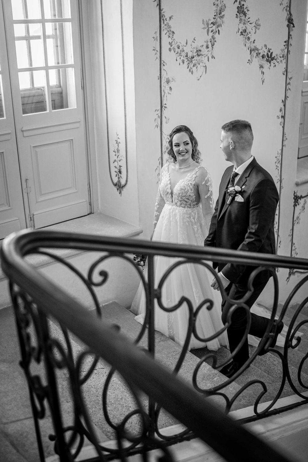 A black and white photograph of a couple in wedding attire standing by an ornate vintage staircase with decorative walls.