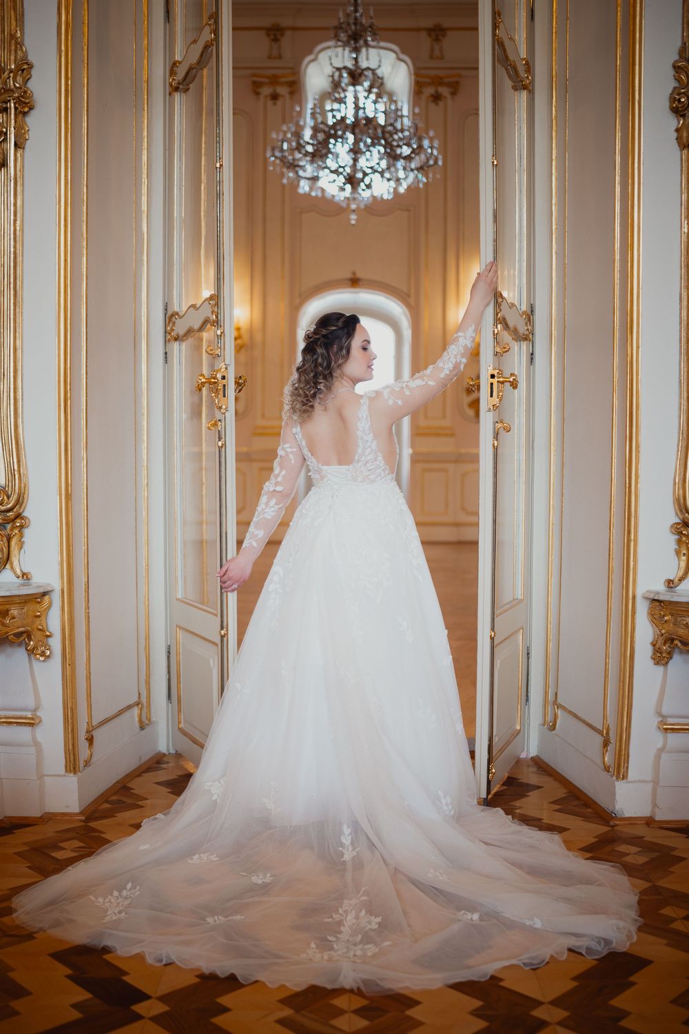 A bridal gown with long lace sleeves and flowing train photographed in an elegant palace room with chandelier and gold details.