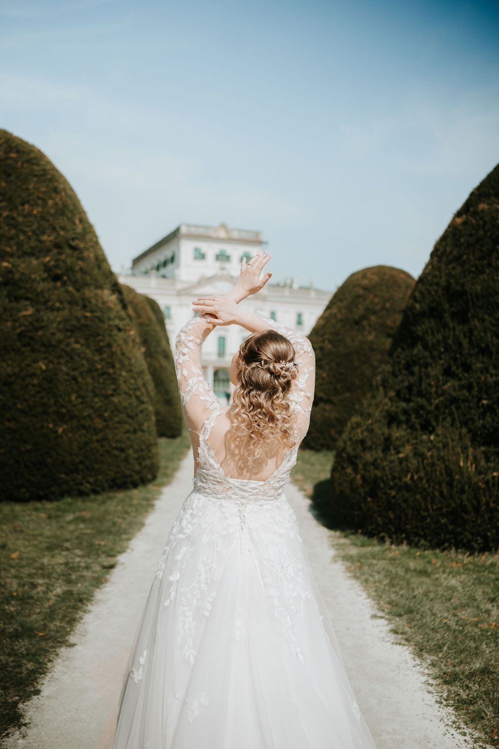 A bride in a lace back wedding dress stands between manicured hedges on a garden path leading to a mansion.