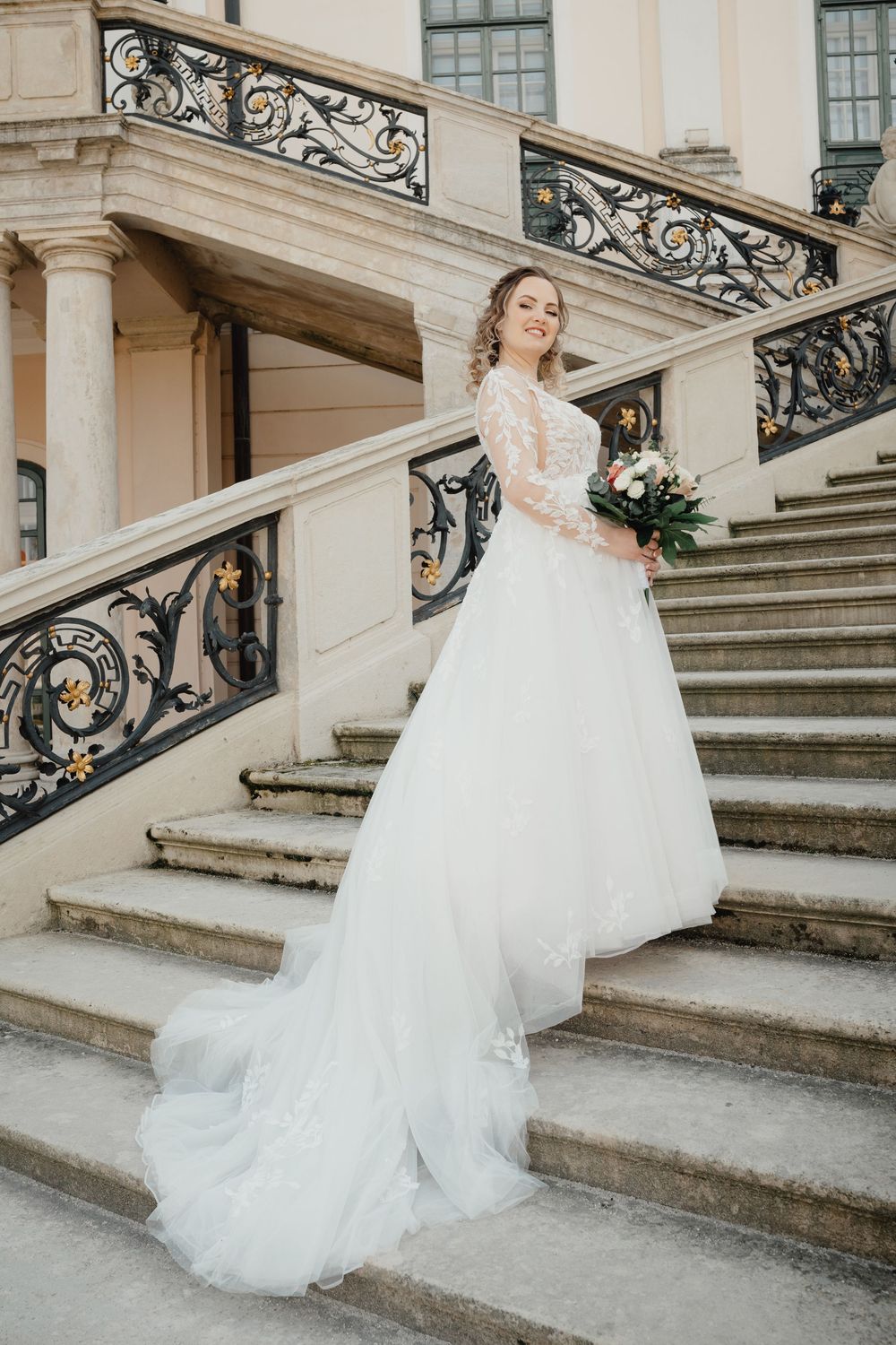 Elegant wedding dress with lace sleeves and flowing tulle skirt photographed on grand stone staircase with ornate railings.