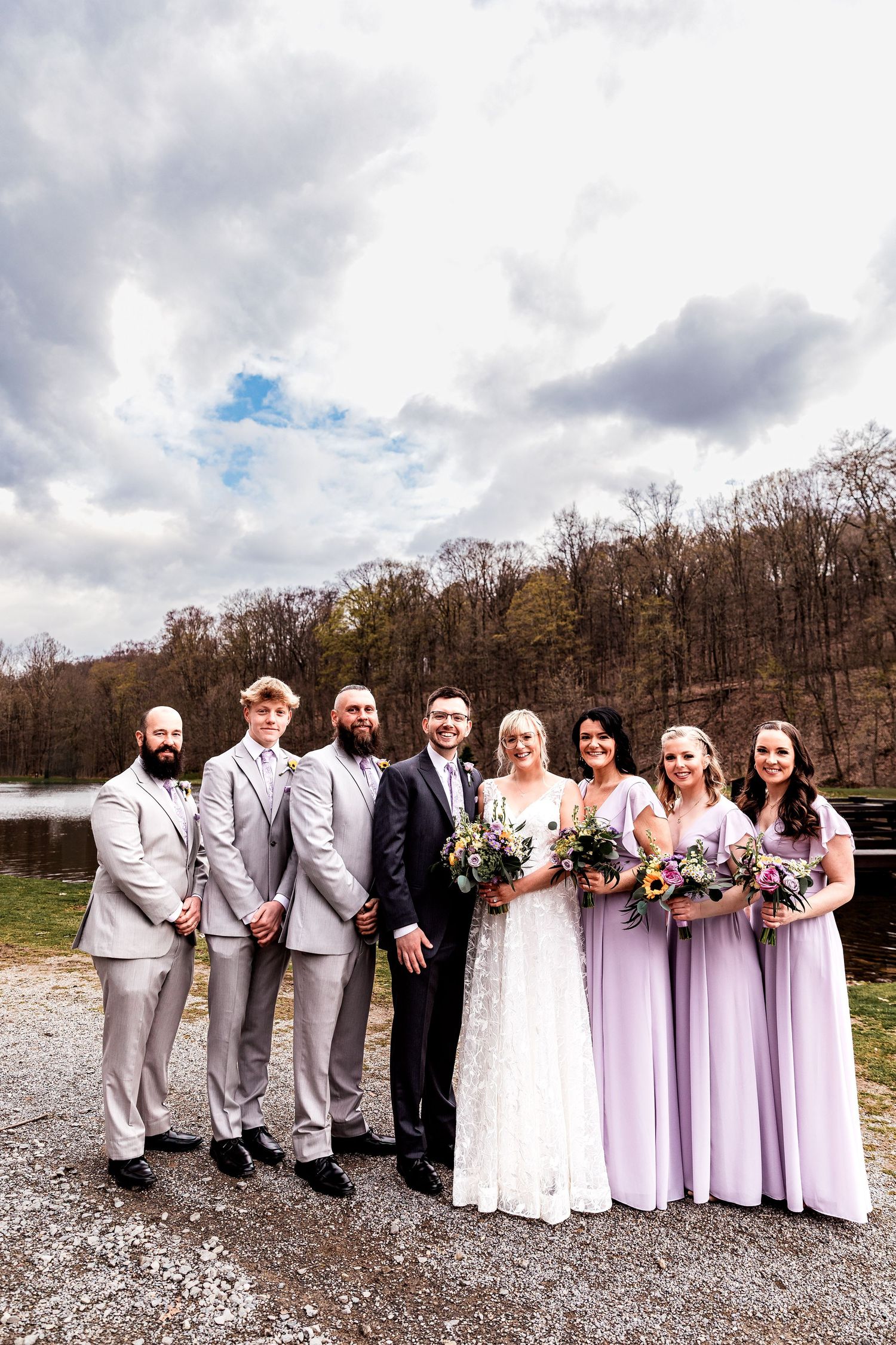 Wedding couple shares kiss while wedding party holds up bottles of craft beer against cloudy sky.
