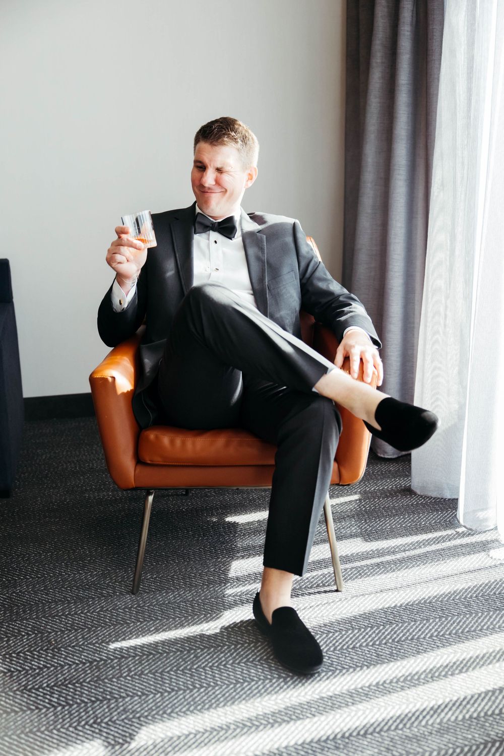Elegant groom in black tuxedo and bow tie sits relaxed in brown leather chair holding champagne glass near window.