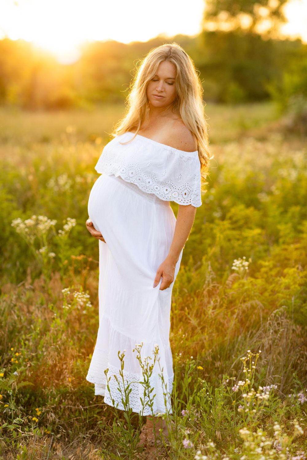 A person in a white off-shoulder dress stands in a golden sunlit meadow during sunset among wildflowers.