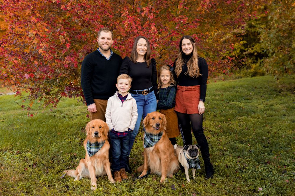 A group portrait on grass with three dogs and vibrant red fall foliage in the background.