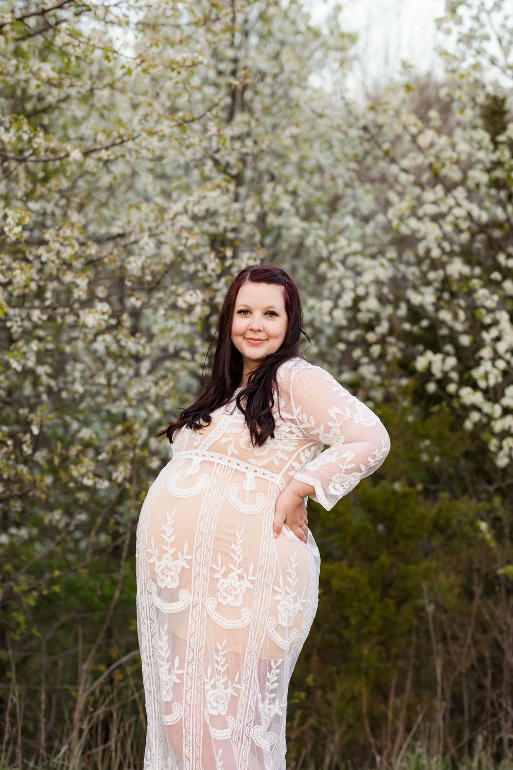 A pregnant figure wearing a sheer lace gown poses in front of blooming spring trees with white blossoms.