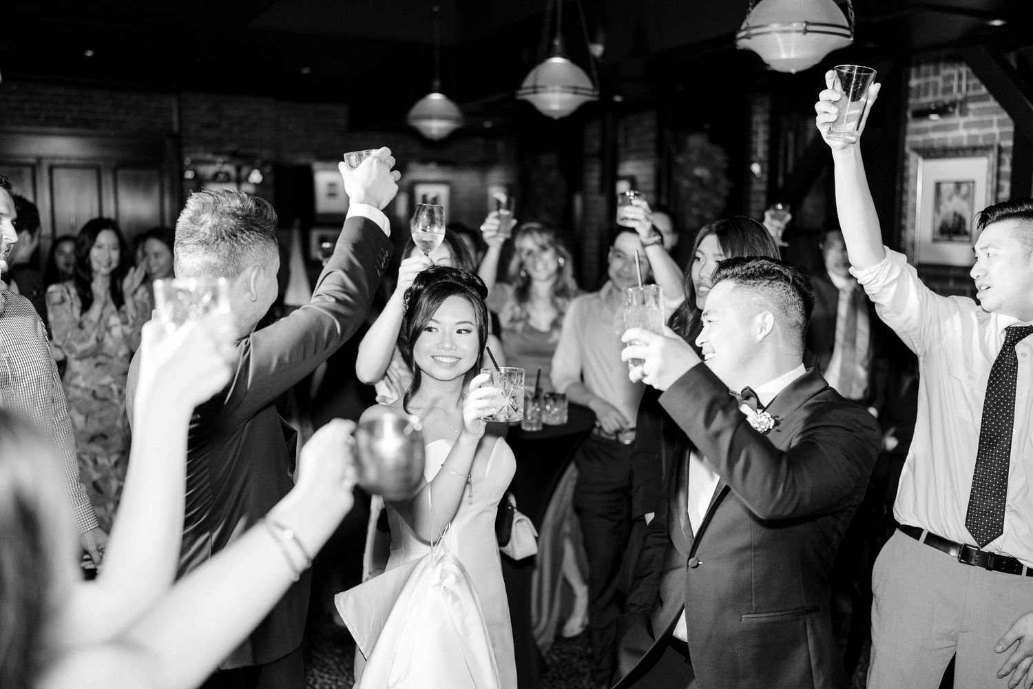 Wedding celebrants dance joyfully at a reception with hands raised high in a dimly lit venue with hanging lights.