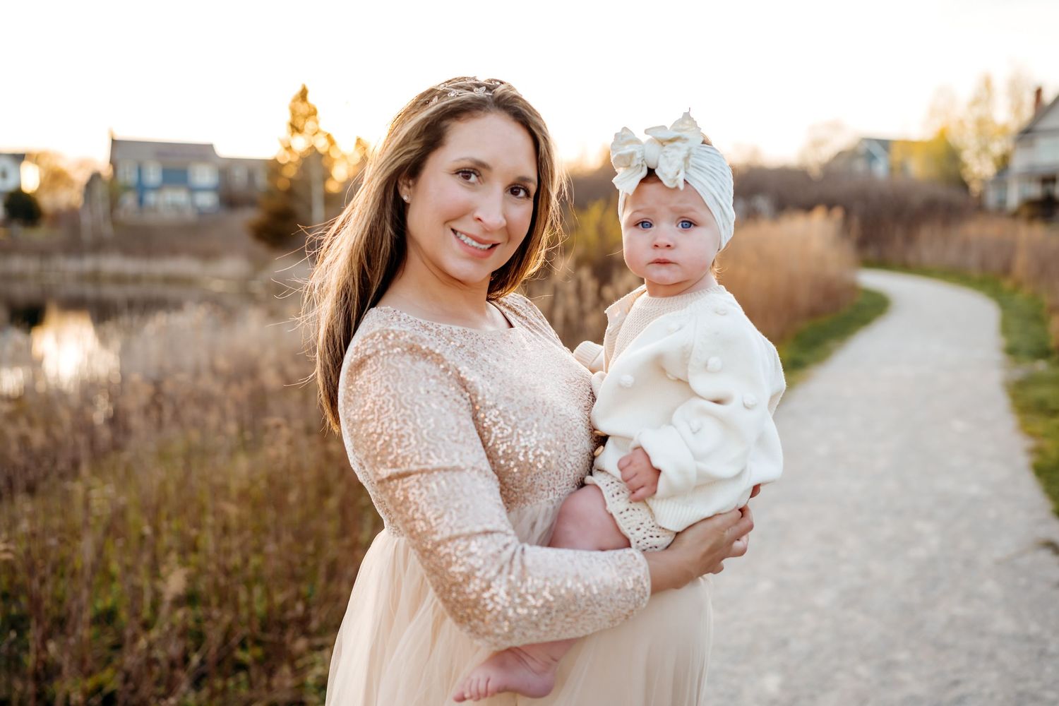 Long-haired person in cream lace dress holds small child wearing white dress and headband on gravel path at sunset.