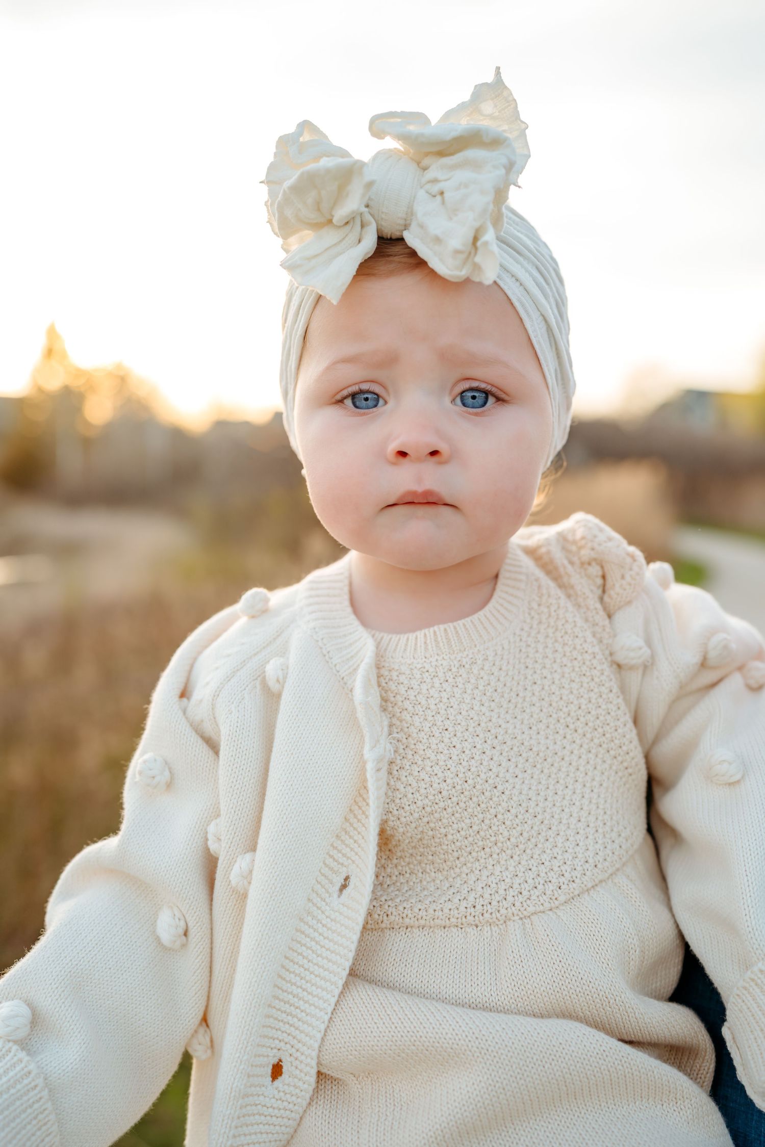 A toddler dressed in cream knit clothing and a bow headband sits outdoors during golden hour sunset.