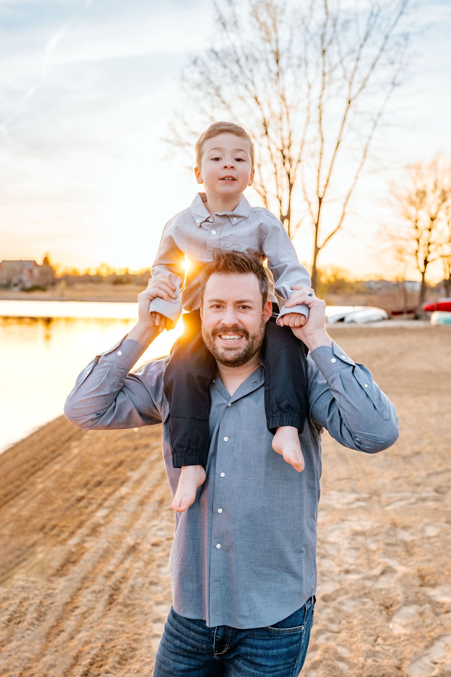 A parent carries a child on shoulders during sunset near a lakeside with bare trees in the background.