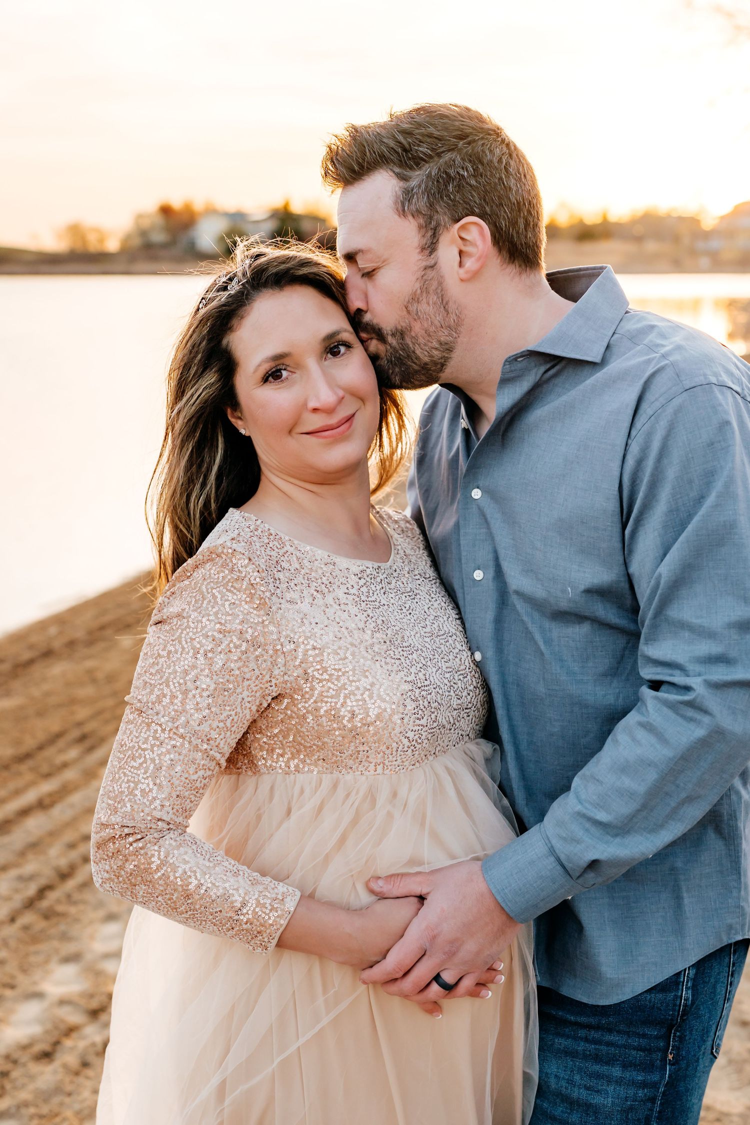 A romantic maternity photo of a couple embracing on a beach at sunset while wearing casual dress clothes.