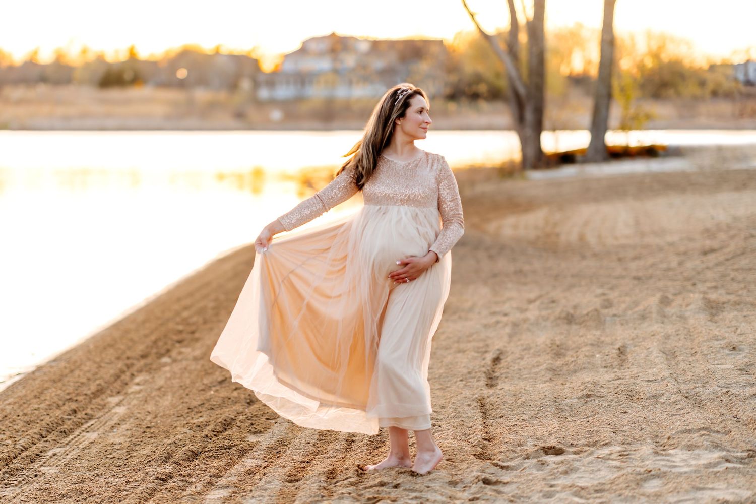 Person in flowing white dress poses gracefully on sandy beach during golden sunset by a lake.