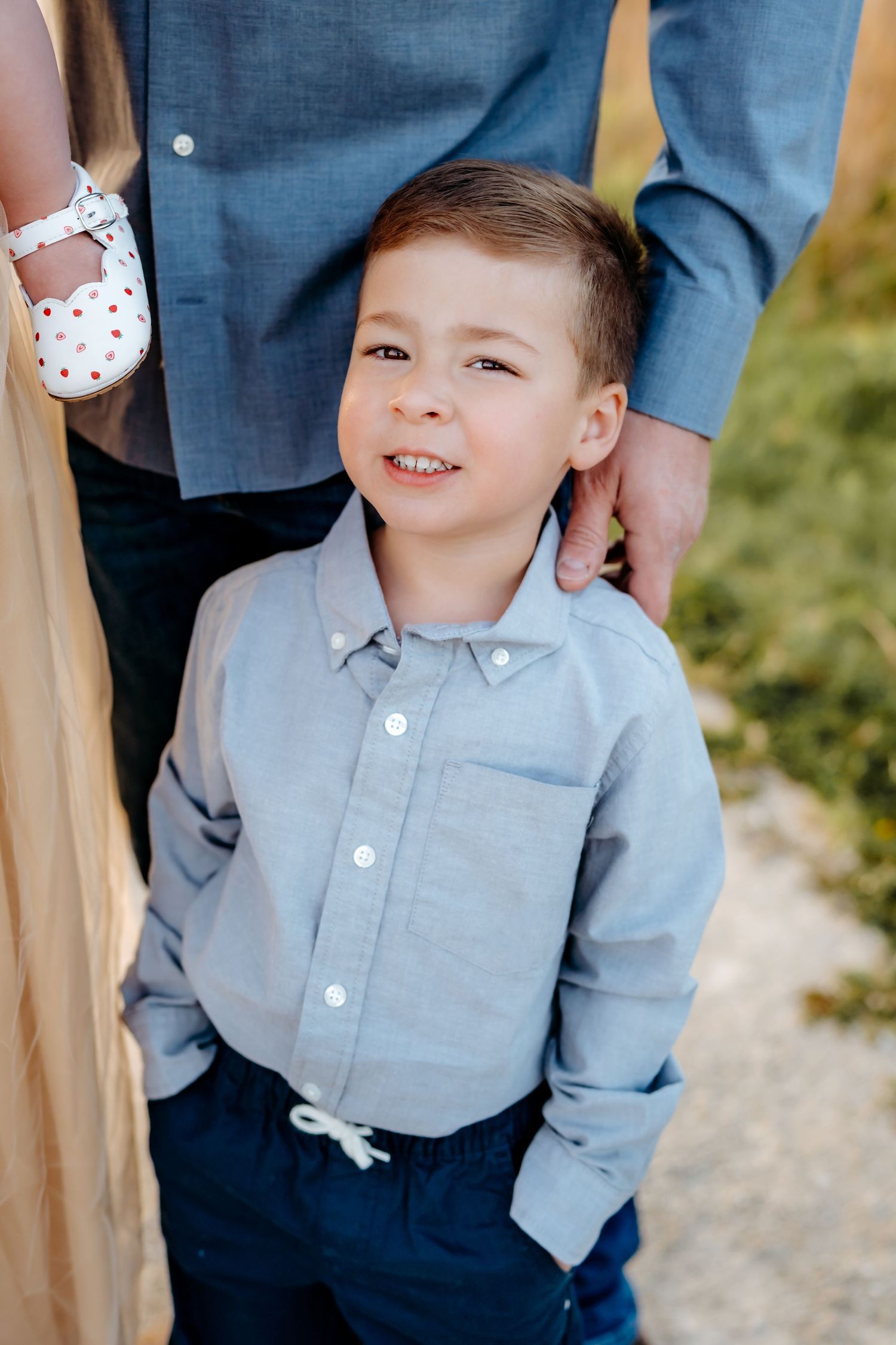A child stands smiling while wearing a light blue button-down shirt and navy pants.