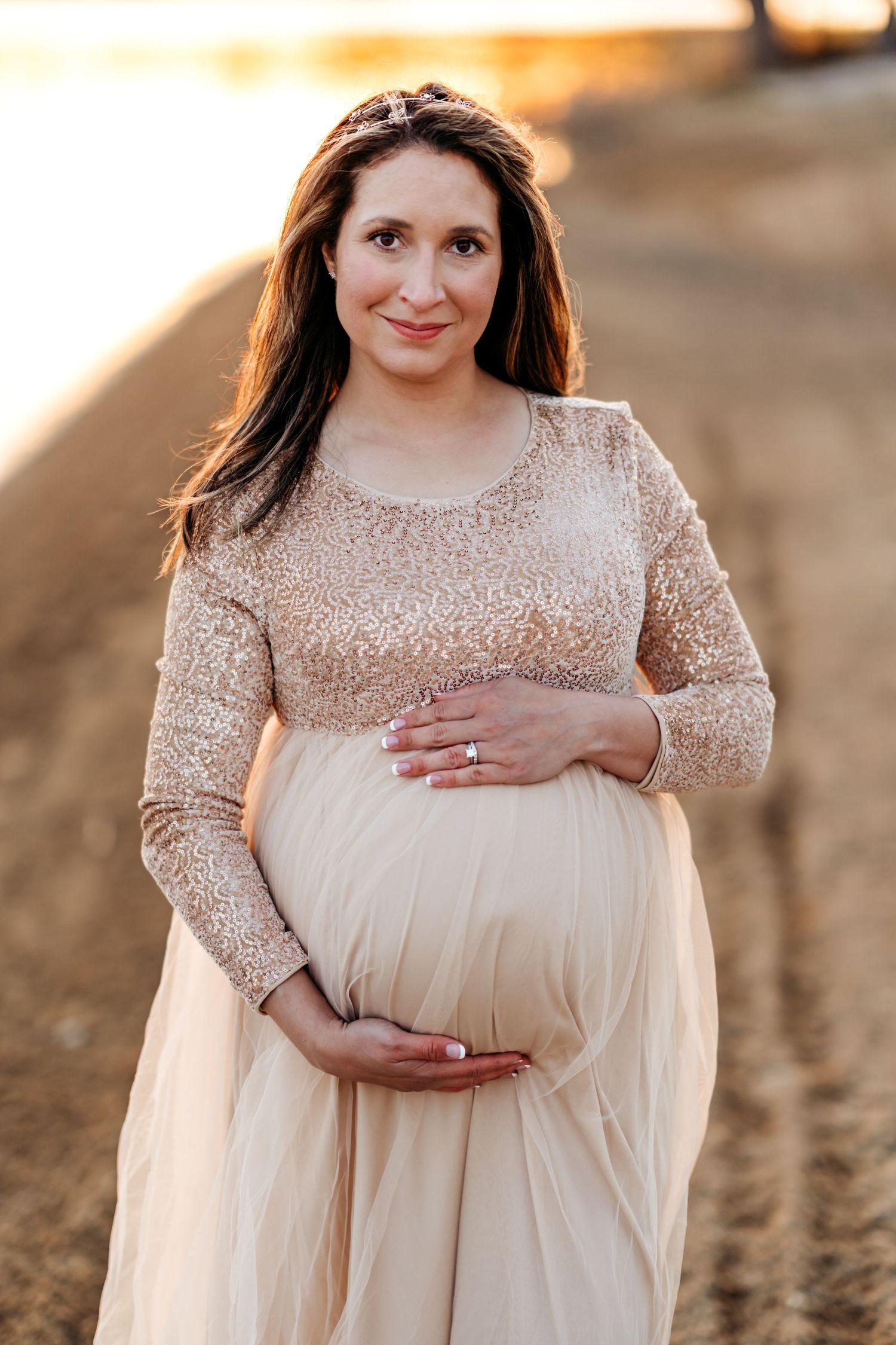Expectant mother in beige lace top and flowy tulle skirt poses outdoors during golden hour sunset.