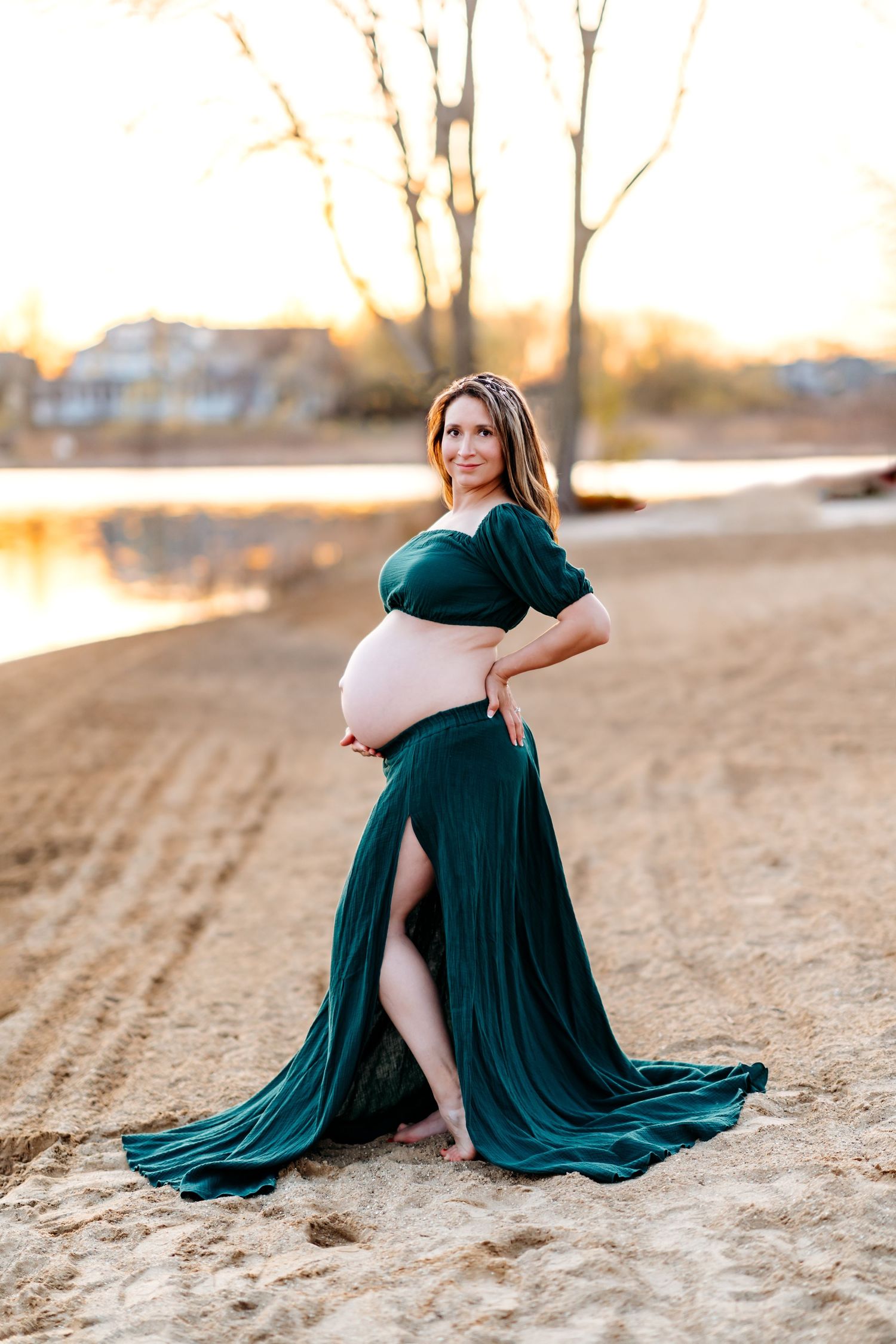 Expectant mother wearing flowing emerald green dress poses gracefully on sandy beach at sunset.