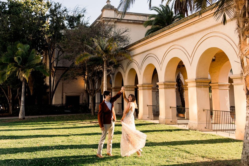 A couple dances in wedding attire on a grassy lawn beside a Spanish colonial building with arched walkways and palm trees.