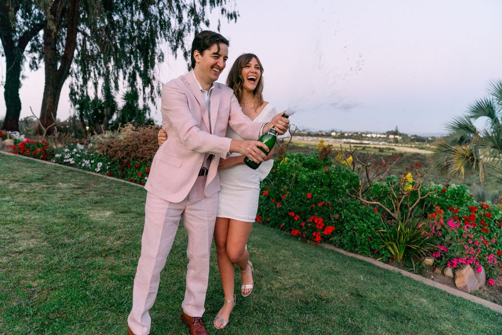 Two people stand on grass by garden with ocean view as one opens bottle of champagne.