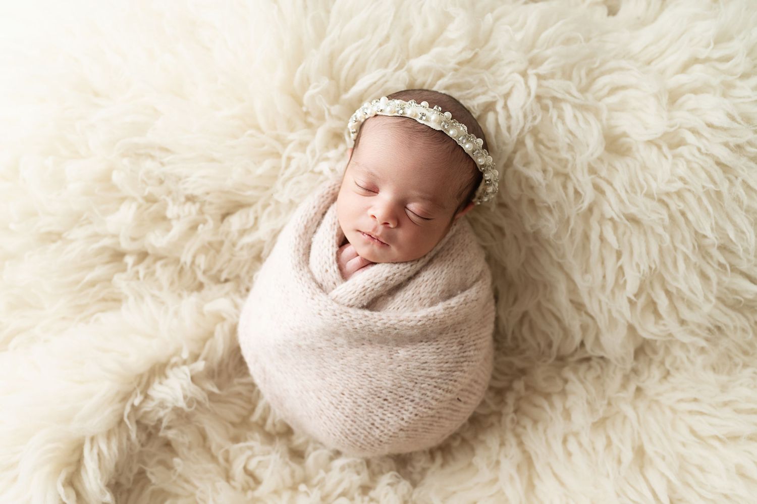Sleeping newborn wrapped in beige swaddle on fluffy white rug.