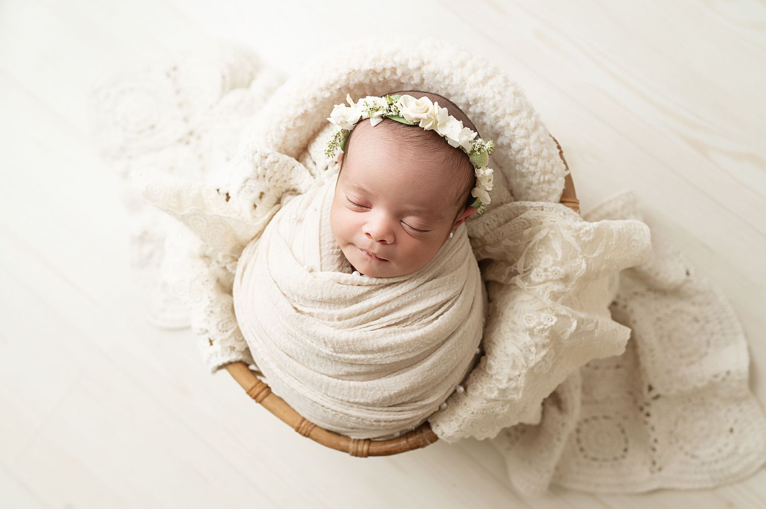 Newborn wrapped in cream swaddle posed in wooden bowl with lace details.