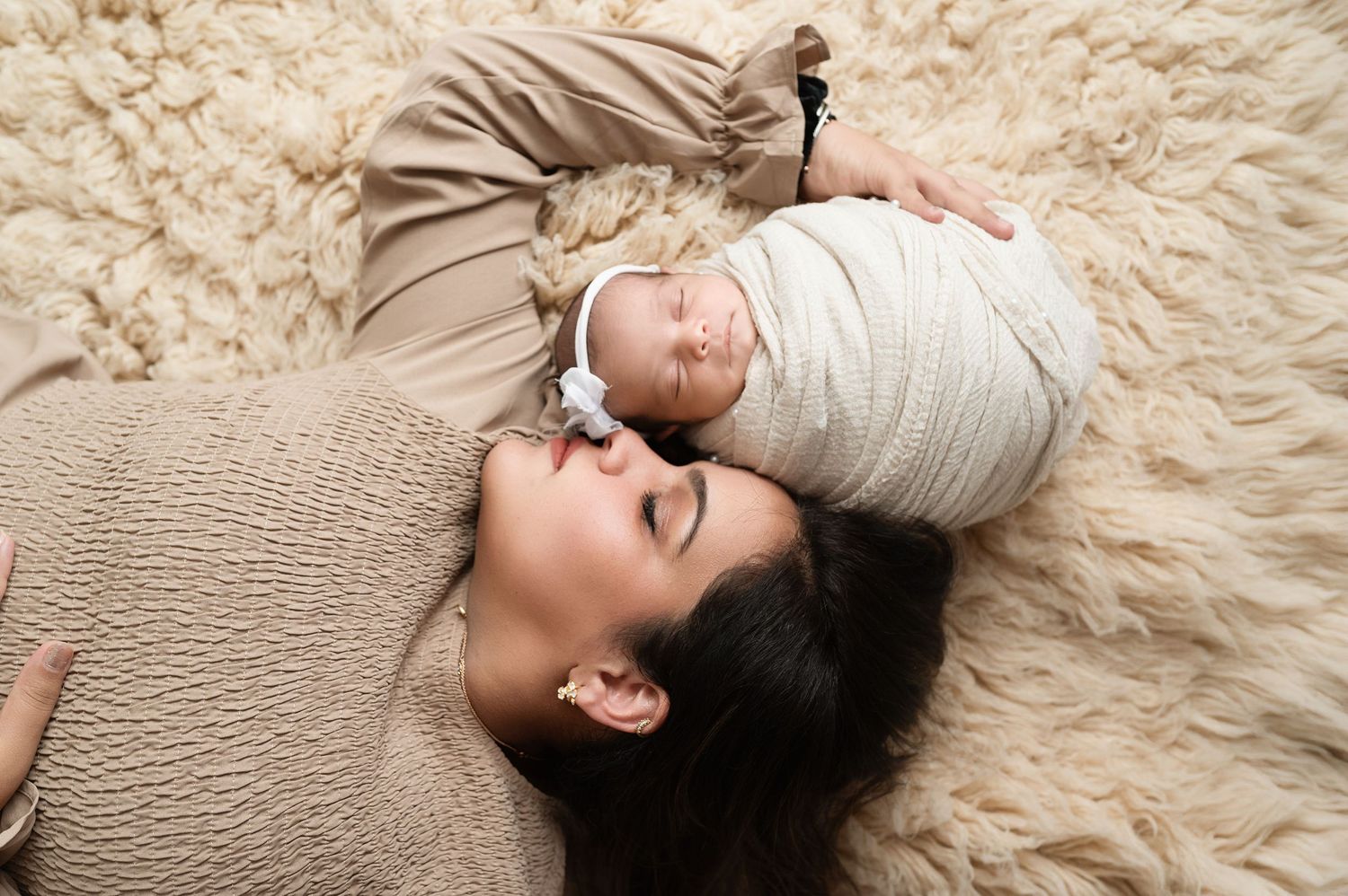 Baby wrapped in beige fabric lies on textured cream fur rug for newborn photography session.