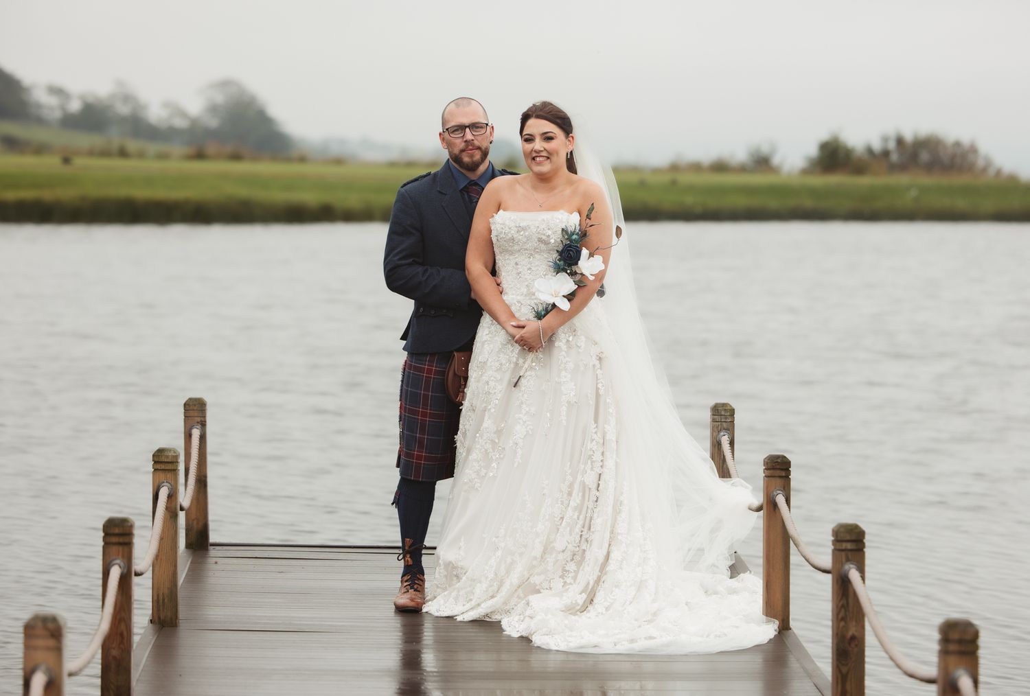 A couple stands together on a wooden dock overlooking a serene lake during a cloudy afternoon.