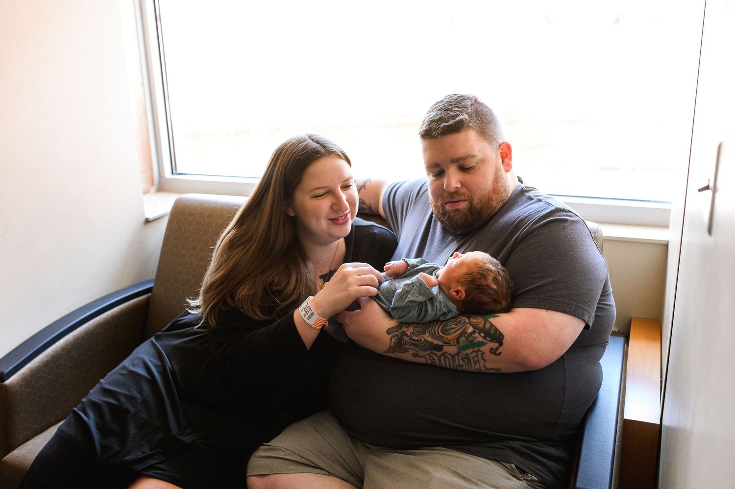 A couple sits together on a couch holding their newborn baby during an intimate indoor photo session.