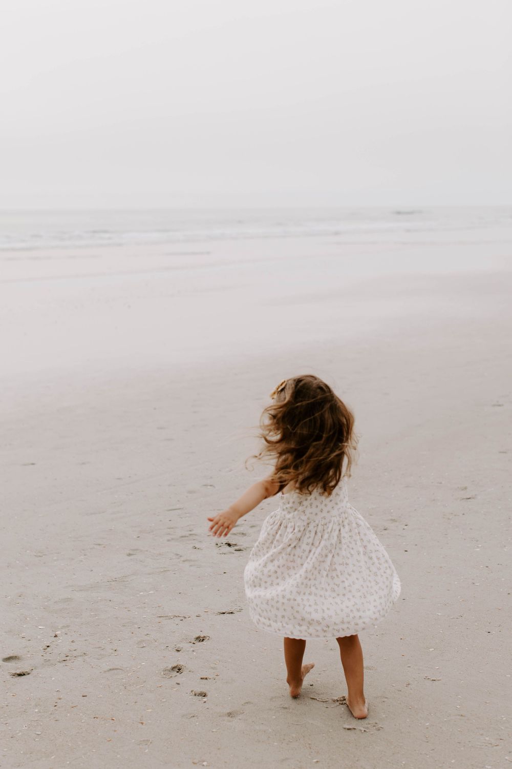 Young child in white dress twirling on empty beach.