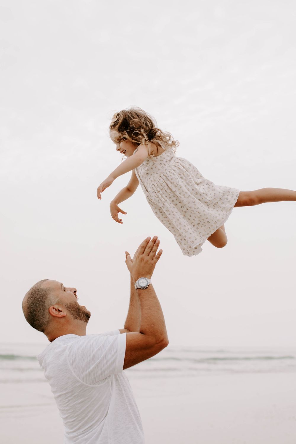 Parent playfully lifts young child in the air at the beach while wearing coordinated outfits.